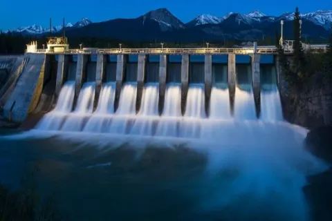 A hydroelectric dam releasing water at dusk, with mountains in the background.